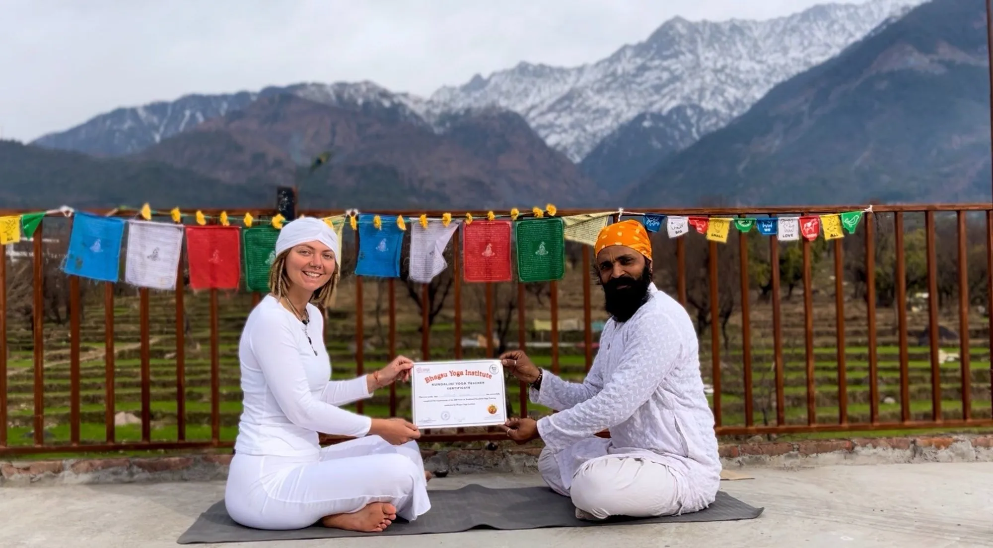 A student and Omji sitting on the terrace holding a certificate, with prayer flags and mountains behind them