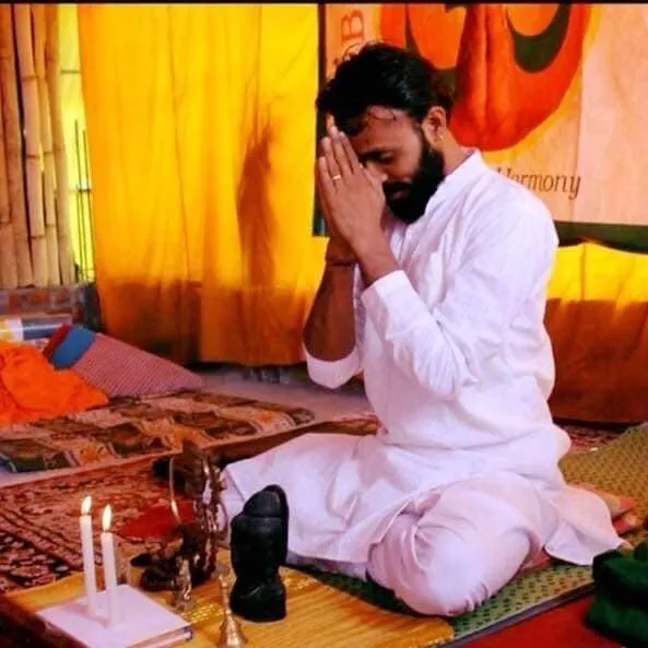 Omji in white clothes sitting cross-legged with folded hands in front of a small altar with candles